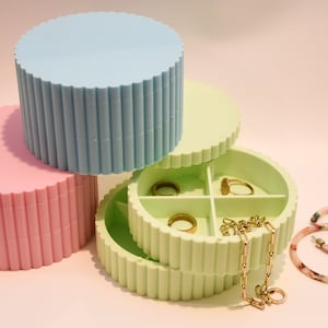 May include: Three round, stackable jewelry boxes in pastel pink, light blue, and light green. The top box has four compartments for rings and a gold chain necklace. The bottom box has a lid with a scalloped edge.