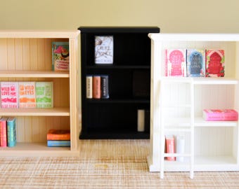 May include: Three miniature bookshelves in natural, black, and white. The shelves are filled with tiny books in various colors, including pink, blue, orange, and green. The white bookshelf has a small ladder attached. The books have titles like "Wild Love" and "Once Upon a Time".