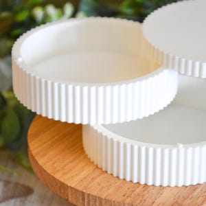 May include: Three white, round, tiered trays with a fluted edge design. The trays are stacked on a round, wooden base. The background features green foliage.