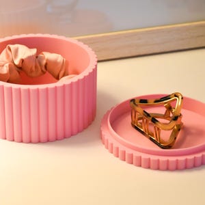 May include: A pink, ribbed, round container with a lid. The container has a beige scrunchie inside. The lid is open and a gold hair clip is sitting on top of the lid.