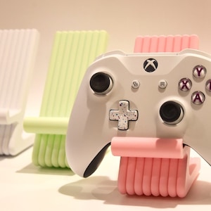 May include: A white Xbox controller rests on a pink stand, with two other stands in the background, one white and one light green. The controller has a speckled cross-shaped directional pad and purple lettered buttons.