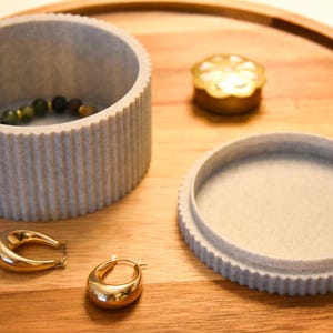 May include: Two gray, textured, round containers with lids, one with a beaded bracelet inside. The containers are on a wooden surface with a gold metal decorative object.