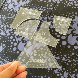May include: Clear acrylic quilting templates labeled A, B, C, D, and E. Each template has a different shape and dotted lines. The text "Popsicle Parade Quilt © 2016 Hello Melly Designs" is printed on the templates. The templates are on a butterfly patterned fabric.