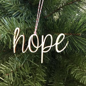 May include: A wooden Christmas ornament in the shape of the word "hope" in a cursive font. The ornament is light brown with a darker brown outline and hangs from a red and white string, set against a backdrop of green Christmas tree branches.