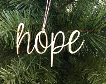 May include: A wooden Christmas ornament in the shape of the word "hope" in a cursive font. The ornament is light brown with a darker brown outline and hangs from a red and white string, set against a backdrop of green Christmas tree branches.