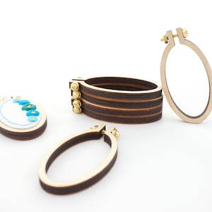 Puede incluir: Aros de bordado de madera y una pequeña pieza bordada con diseños florales azules y turquesas. Los aros son ovalados, con herrajes de latón. Se ve una pila de aros, junto con dos aros individuales.