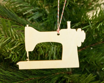 May include: A light brown wooden sewing machine Christmas tree ornament, suspended by a red and white string. The ornament is set against a backdrop of green fir tree branches. The sewing machine is a classic design.