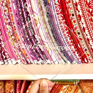 May include: A clear acrylic Pillow Corner Template is held in front of a shelf of colorful fabric. The fabric is arranged in vertical stacks, showcasing various patterns in shades of pink, purple, and red. The template has the words "Pillow Corner Template" printed on it.