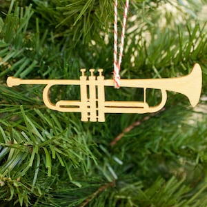 Op de afbeelding: Een houten trompet kerstversiering hangend aan een groene kerstboom. De versiering is lichtbruin en heeft een rood-wit touwtje om op te hangen. De trompet is een gedetailleerde uitsparing van het instrument.