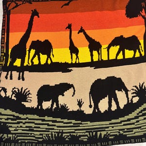 Può includere: Una coperta rettangolare all'uncinetto che raffigura un paesaggio di savana con sagome nere di giraffe ed elefanti su un gradiente di tramonto giallo, arancione e rosso. La sezione inferiore mostra un terreno color marrone chiaro.