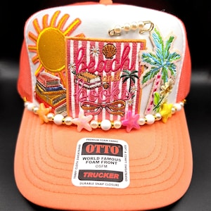 May include: A coral-colored trucker hat with a white front panel featuring embroidered beach-themed designs. The design includes a sun, palm trees, books, and the words "beach and books." The brim is adorned with pearls and star-shaped embellishments.