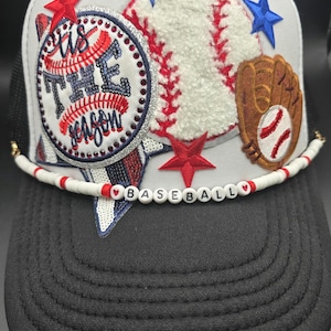 May include: White trucker hat with a baseball theme. It features a red, white, and blue baseball, a brown baseball glove, a red star, and a blue star. The hat also has a white beaded strap with the words "BASEBALL" in black letters.