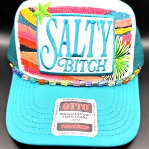 May include: A turquoise and white trucker hat featuring a rectangular patch with the text SALTY BITCH in blue embroidery. The design includes a green star, a palm leaf, and colorful beaded trim. An OTTO brand sticker is on the turquoise brim.