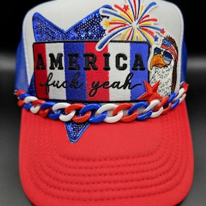 May include: A red, white, and blue trucker hat with the text "AMERICA fuck yeah." The hat features a sequined blue star, a cartoon eagle wearing sunglasses, and a chain around the brim. A fireworks graphic is also present.