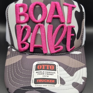 May include: A trucker hat with a camouflage pattern in shades of gray, white, and black. The hat features the embroidered text "BOAT BABE" in bright pink. The hat has a white mesh back and a snap closure.