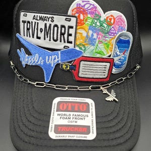 May include: Black trucker hat with a mesh back and a curved brim. The front features embroidered patches with travel-themed designs, including a license plate that reads "ALWAYS TRVL MORE" and a blue airplane. A silver chain and airplane charm are attached.