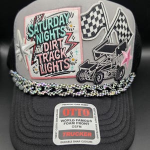 May include: A grey and black trucker hat featuring a patch with the text SATURDAY NIGHTS AND DIRT TRACK LIGHTS, a sprint car graphic, and two checkered flags. A metallic chain trim sits above the brim, which bears an OTTO brand sticker.