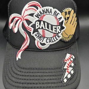 May include: Black baseball cap with a mesh back and a curved brim. The cap features embroidered patches including a baseball, a baseball glove, a lightning bolt, and the text "WANNA BE A BALLER SHOT CALLER".