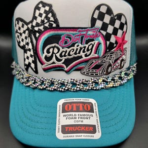 May include: A teal and white trucker hat featuring a checkered bow patch, a heart-shaped checkered patch, a pink star, and a Dirt Track Racing patch. A metallic checkered chain adorns the base of the crown. An Otto brand sticker is on the brim.