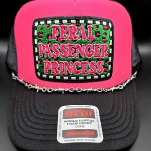 May include: A hot pink and black trucker hat featuring a rectangular patch with a green leopard print background and the embroidered pink text FERAL PASSENGER PRINCESS. A silver-toned barbed wire chain accents the base of the front foam panel.