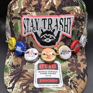 Gorra de camionero Stay Trashy con mapache y cadena de chapa de cerveza sobre un nuevo estampado de camuflaje.