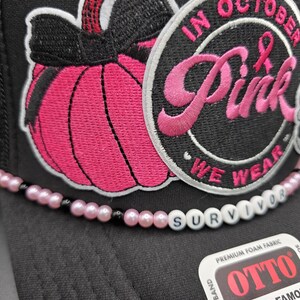 May include: Black trucker hat with a pink pumpkin patch and the words "IN OCTOBER Pink WE WEAR" embroidered on the front. A beaded strand spells "SURVIVOR" across the brim. The hat is made of premium foam fabric.