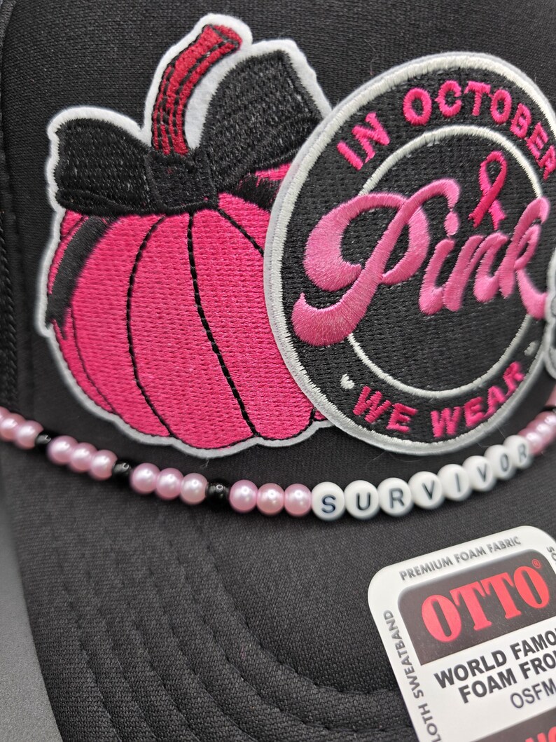 May include: Black trucker hat with a pink pumpkin patch and the words "IN OCTOBER Pink WE WEAR" embroidered on the front. A beaded strand spells "SURVIVOR" across the brim. The hat is made of premium foam fabric.