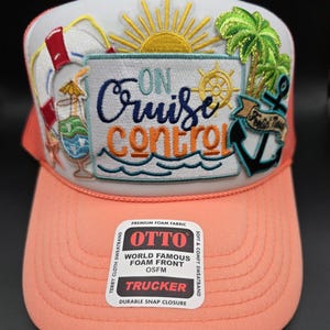 May include: A coral and white trucker hat with the phrase "On Cruise Control" embroidered on the front. The design includes a sun, palm tree, anchor, and a cocktail. The hat has a mesh back and a snap closure.