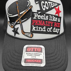 May include: Black and gray trucker hat with a hockey puck and crossed hockey sticks graphic. The hat features the text "GO TEAM" and "Feels like a PENALTY BOX kind of day". The hat has a snap closure.