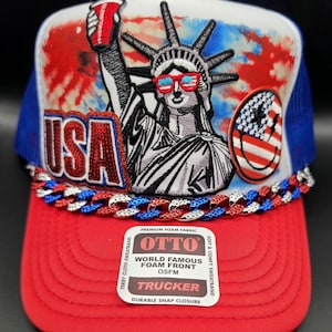 May include: A red, white, and blue trucker hat featuring an embroidered Statue of Liberty wearing flag-patterned sunglasses and holding a red cup. The hat includes a red, white, and blue chain across the brim and a sticker reading OTTO.