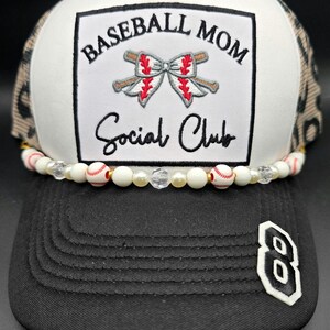 May include: A baseball cap with a black bill and a white and leopard print mesh back. The front features a white patch with the words "Baseball Mom Social Club" and baseball bat and bow graphic. A beaded strand with baseballs and pearls adorns the bill.