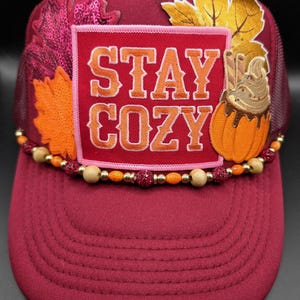 May include: A maroon trucker hat featuring a rectangular patch with the text STAY COZY in orange and white. The hat is decorated with sequined autumn leaf appliques, a pumpkin graphic, and a string of wooden, orange, and glittery beads.