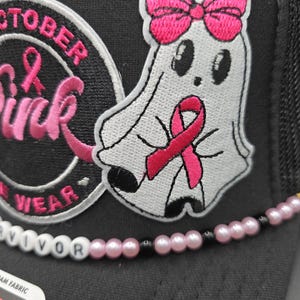 May include: Black trucker hat with a white ghost patch wearing a pink ribbon and bow. The hat has the words "October Pink" and "Survivor" on it. The hat has a pink and black beaded accent.
