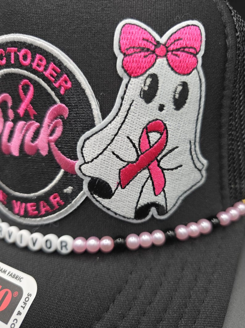 May include: Black trucker hat with a white ghost patch wearing a pink ribbon and bow. The hat has the words "October Pink" and "Survivor" on it. The hat has a pink and black beaded accent.