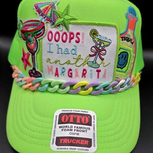 May include: A neon green trucker hat featuring a central patch with the text OOOPS! I had another MARGARITA, surrounded by embroidered cocktail and tequila bottle motifs. A pastel-colored plastic chain trim sits above the brim, which has an OTTO brand sticker.