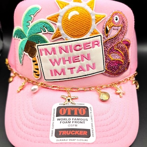 May include: A light pink trucker hat featuring embroidered patches of a palm tree, a yellow sun, and a pink flamingo. A rectangular patch reads "I'M NICER WHEN IM TAN" in pink text. A gold chain with starfish, seashell, and pearl charms hangs across the front.