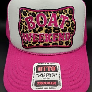 May include: A pink trucker hat with a white front panel and pink mesh backing. The hat features the words "BOAT WEEKEND" in pink letters on a leopard print patch. The hat also has a "OTTO" label.