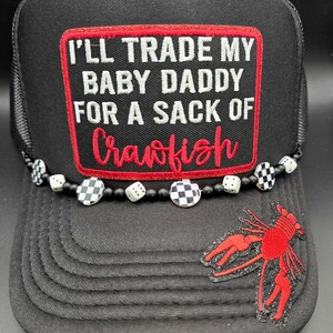 May include: Black trucker hat with a mesh back and a curved brim. The front patch reads "I'll Trade My Baby Daddy For A Sack Of Crawfish" in white and red. A red crawfish patch and a beaded dice accent the hat.