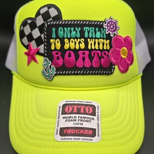 May include: A neon green trucker hat with a white mesh back. The front of the hat features a black and white checkered heart, a red star, a blue anchor, and a pink flower. The hat also has a patch that says "I only talk to boys with boats". The hat is made by Otto and has a foam front, a terry cloth sweatband, and a durable snap closure.
