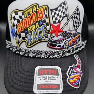 May include: A black and white trucker hat featuring an embroidered patch with the text IT'S A GOOD DAY FOR A RACE, checkered flags, a red star, and a race car. A black and white checkered chain adorns the brim, alongside a fuel can patch and an OTTO label.