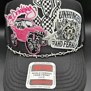 May include: Black trucker hat with a mesh back and a curved brim. The front features embroidered patches including a pink off-road vehicle, a raccoon, and the words "Day Drinking" and "Unhinged and Feral". A barbed wire design accents the brim.