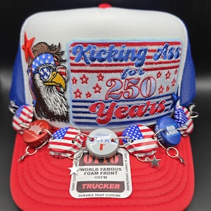 May include: A white and red trucker hat featuring an embroidered patch with an eagle wearing star-spangled sunglasses and the text Kicking Ass for 250 Years. The brim is red, and the hat is decorated with various metal bottle caps and small stars.