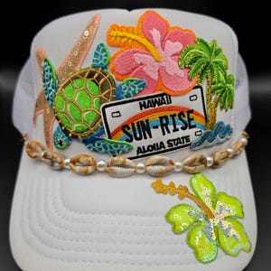 May include: A white trucker hat featuring embroidered patches of a green sea turtle, a pink hibiscus flower, a palm tree, a starfish, and a Hawaii license plate reading SUN-RISE ALOHA STATE. A string of cowrie shells and white beads accents the brim.