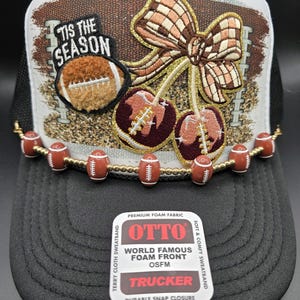 May include: A black trucker hat with a white and brown front panel featuring a football-themed design. The design includes a football, a bow, and cherries. The hat has a beaded football embellishment along the brim and the text "TIS THE SEASON".