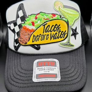 May include: A black and white trucker hat with a white front panel featuring an embroidered taco, margarita, and the text "Tacos before vatos." The hat has a black mesh back and a curved brim.