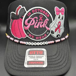 May include: Black trucker hat with a pink and white embroidered design. The design includes a pink pumpkin, a ghost with a pink ribbon, and the words "In October We Wear Pink". The word "Survivor" is spelled out in white beads across the brim.