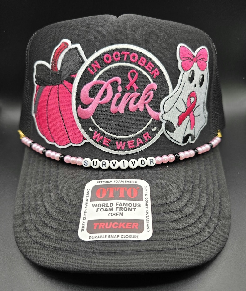 May include: Black trucker hat with a pink and white embroidered design. The design includes a pink pumpkin, a ghost with a pink ribbon, and the words "In October We Wear Pink". The word "Survivor" is spelled out in white beads across the brim.