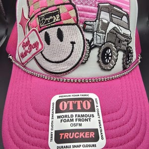 May include: Pink trucker hat with a white mesh back. The front of the hat features a pink and white checkered beanie, a smiley face, a pink and white American flag, and a gray ATV. The hat has a white label with the text "OTTO", "WORLD FAMOUS FOAM FRONT", "OSFM", "TRUCKER", and "DURABLE SNAP CLOSURE".