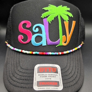 May include: Black trucker hat with the word "Salty" in colorful embroidered letters and a green palm tree. The hat has a mesh back, a beaded band, and a "World Famous Foam Front" tag.