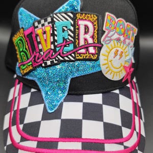 May include: A black baseball cap with a checkered brim featuring two pink stripes. The front displays a large blue sequined star patch with the text RIVER BRAT in colorful letters, alongside a patch with the text BOAT BABY and a yellow sun.